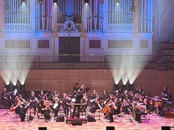 LEAP orchestra is performing on a stage illuminated by spotlights, with a large organ visible in the background.